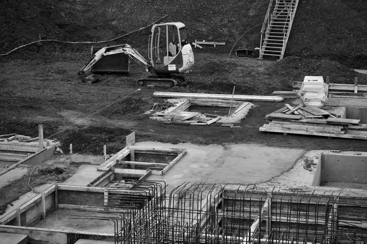 services-02 Monochrome image of an excavator at a construction site with foundations laid and rebar in place.